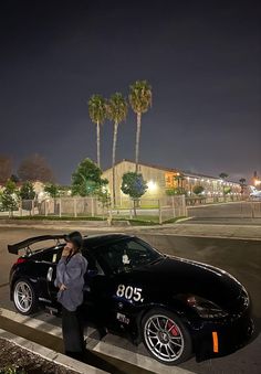 a woman standing next to a black sports car in the parking lot at night with palm trees