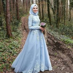 a woman in a blue dress standing on a dirt road with trees and bushes behind her