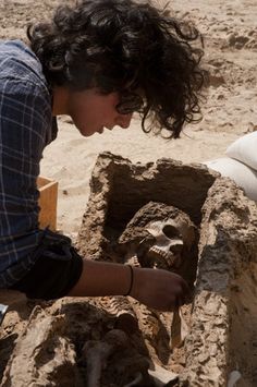 a man kneeling down next to a sand sculpture on the beach with a skull in it's mouth