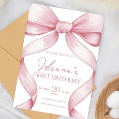 a pink ribbon and bow birthday party card on top of a wooden plate next to cotton balls