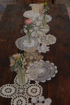 several vases with flowers are lined up on a long table, along with doily