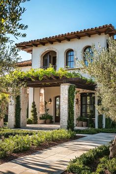 a white house with lots of windows and plants on the front porch, surrounded by greenery