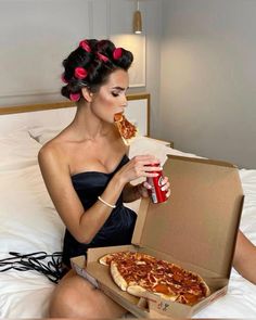 a woman sitting on a bed eating pizza and holding a can of soda in her hand