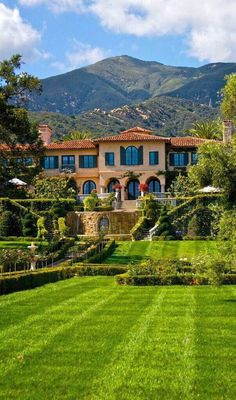 a large house surrounded by lush green trees and bushes with mountains in the background on a sunny day