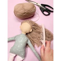 a hand holding a doll next to a pair of scissors and yarn on a pink surface