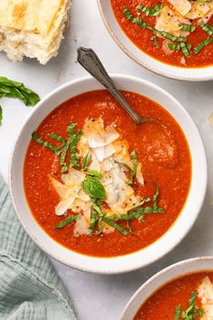 three bowls of tomato soup with bread on the side and basil garnishes