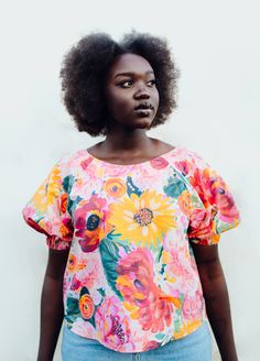 a woman with an afro standing in front of a white wall wearing a colorful top