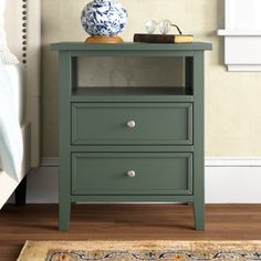 a night stand with two drawers and a blue vase on the top, next to a bed