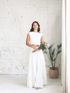 a woman standing in front of a white brick wall holding a bouquet of flowers and smiling at the camera