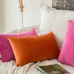 four different colored pillows sitting on top of a bed next to a book and lamp