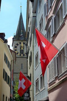 a clock tower is in the distance between two buildings with flags flying from it's sides