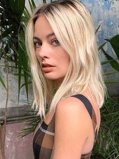 a woman with blonde hair is standing in front of some plants and looking at the camera