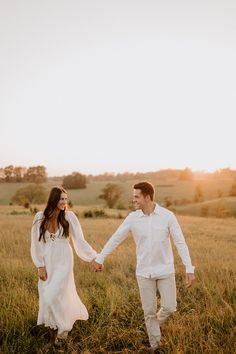 a man and woman holding hands walking through the grass in an open field at sunset