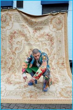 a man kneeling down in front of a large rug on the ground next to a blue building