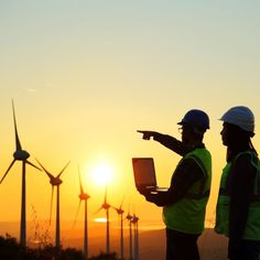 two people in hard hats and vests are looking at windmills as the sun sets