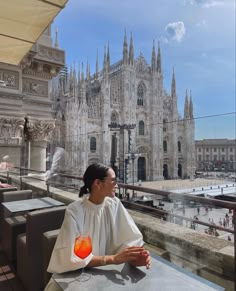 a woman sitting at a table with a drink in front of a large building that looks like a cathedral