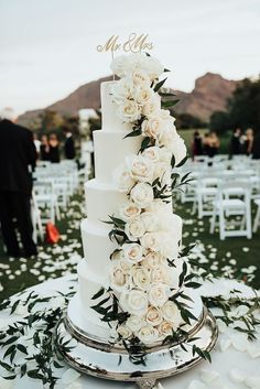 a wedding cake with white flowers and greenery sits on a table in the middle of an outdoor ceremony