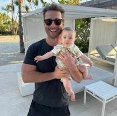 a man holding a baby in his arms while standing next to a white beach chair