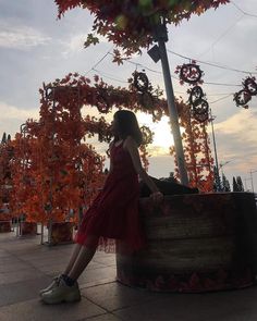 a woman in a red dress is sitting on a barrel near trees with orange leaves