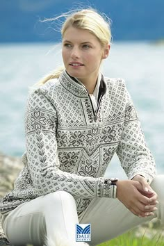 a woman is sitting on the grass by the water wearing white pants and a patterned jacket