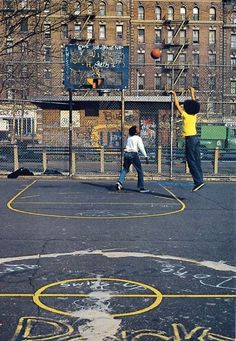 two young boys playing basketball on an outdoor court with graffiti written on the ground and buildings in the background