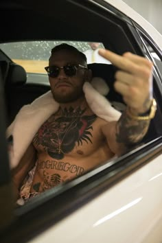 a shirtless man with tattoos sitting in a car pointing to the side while wearing sunglasses