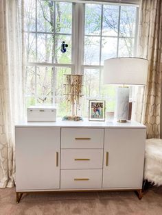 a white sideboard with gold handles and drawers in front of a large window next to a bed