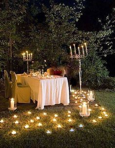 an outdoor dinner table with candles on the grass in front of trees and bushes at night