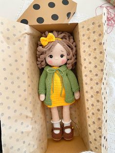 a small doll in a box with polka dots on the wall behind it is wearing a green jacket and yellow dress