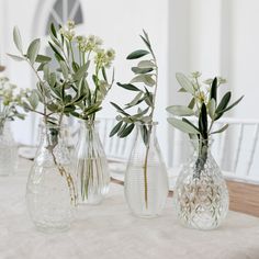 three clear vases with flowers and greenery in them on a white table cloth