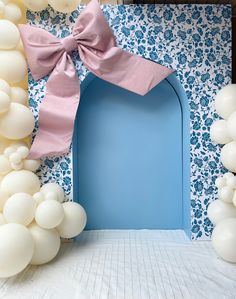 a blue door surrounded by white balloons and pink bow on the top, in front of a floral wallpapered backdrop