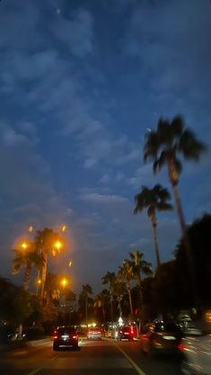 cars driving down the street at night with palm trees in the foreground and lights on