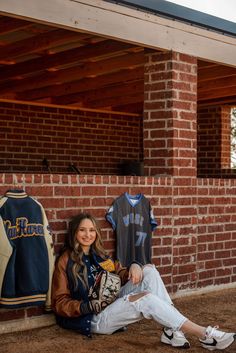 a woman sitting on the ground in front of a brick wall with two baseball jerseys