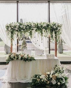 a table with white flowers and greenery on it in front of a large window