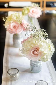 two vases with flowers are sitting on a long white table cloth, along with wedding rings