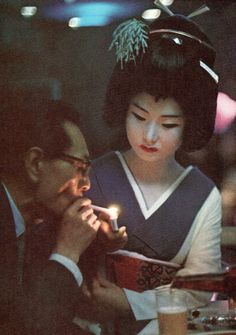 a man and woman sitting at a table with a lit candle in their hands, both wearing geisha garb