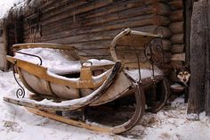 an old sleigh sitting next to a log cabin in the snow with a dog looking at it