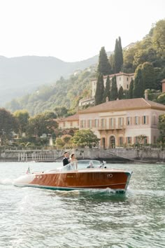 two people in a motorboat on the water near a large house and trees with mountains in the background