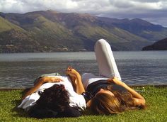 two people laying on the grass near water with mountains in the background and one person reading a book