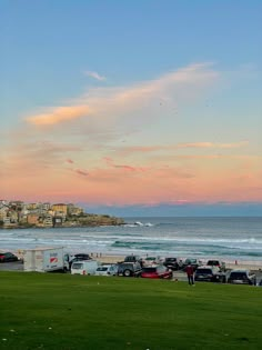 cars are parked on the grass next to the beach as the sun is setting in the distance