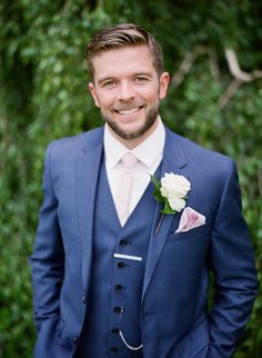 a man in a blue suit and pink tie smiles at the camera while wearing a white rose boutonniere