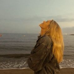 a woman standing on top of a beach next to the ocean looking up at a kite flying in the sky