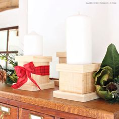 two white candles sitting on top of a wooden table next to greenery and pine cones