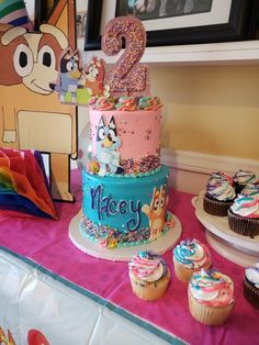 a colorful birthday cake and cupcakes on a table with a pink tablecloth