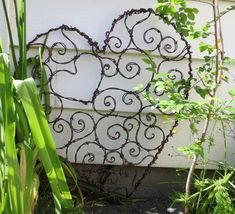 a heart shaped iron sculpture sitting in the grass next to some plants and flowers outside