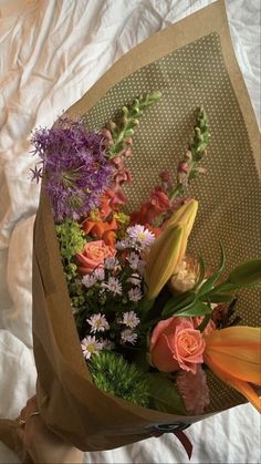 a bouquet of flowers sitting on top of a white sheet covered bed next to a brown paper bag