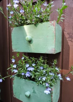 two wooden boxes with flowers in them on the side of a fence, one is painted green and the other is blue