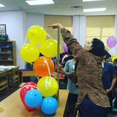 a woman pointing at balloons on a table in a room with other people standing around