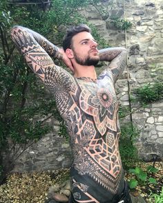 a man with tattoos on his body sitting in front of a stone wall and holding his hands behind his head