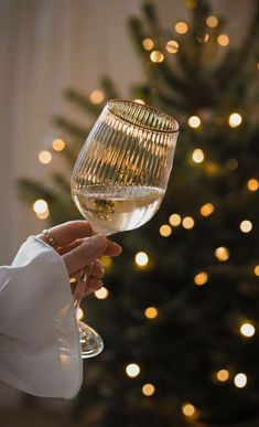 a person holding a wine glass in front of a christmas tree with lights on it